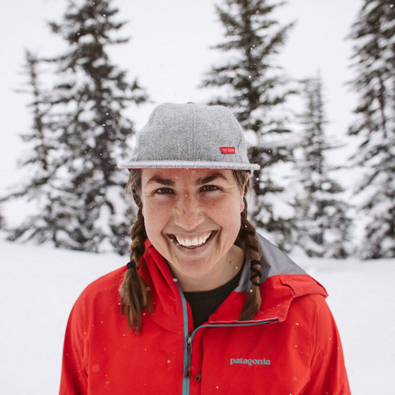 The image shows a woman in a snowy, forested environment. She is wearing a red Patagonia jacket and a grey cap, and her hair is styled in braids. She is smiling at the camera. The background features snow-covered trees, suggesting a winter setting. The overall impression is one of outdoor activity and a cheerful disposition in a cold climate.